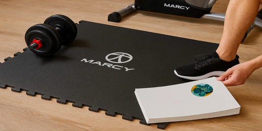 A black Marcy EVA foam fitness mat with logo lies in a home gym, holding a dumbbell and shoe, with a recumbent bike blurred in the background.