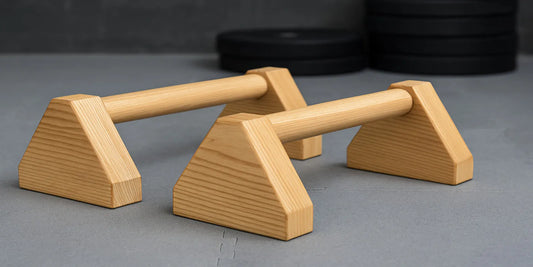 Pair of wooden mini parallettes on a gray gym floor, designed for calisthenics, pushups, and strength training, with weights in background.