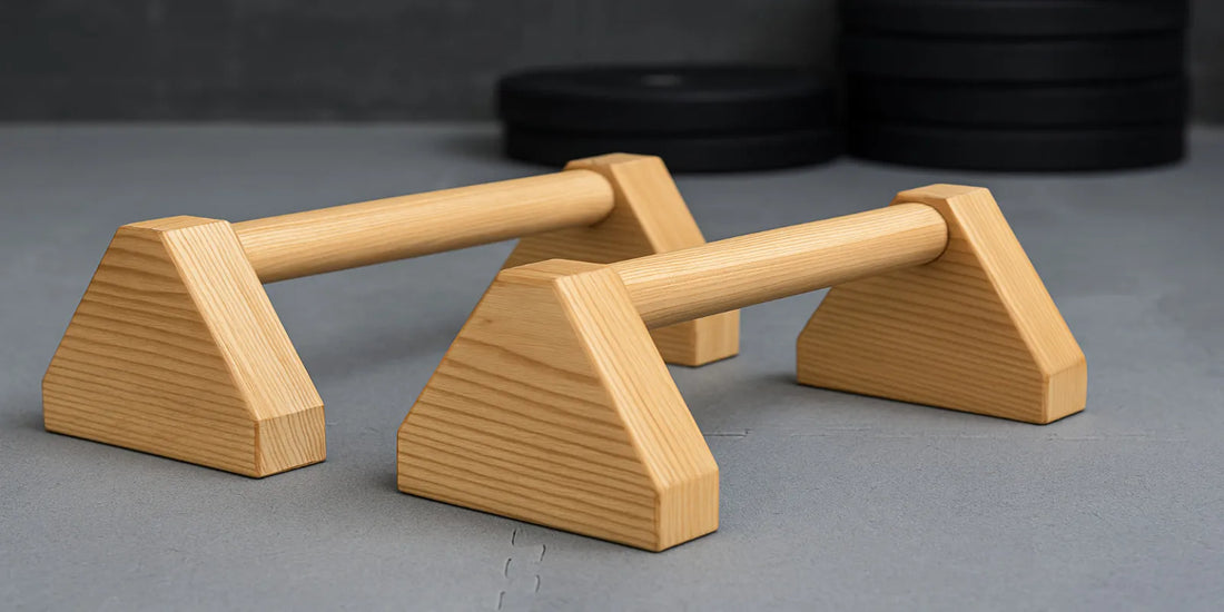 Pair of wooden mini parallettes on a gray gym floor, designed for calisthenics, pushups, and strength training, with weights in background.