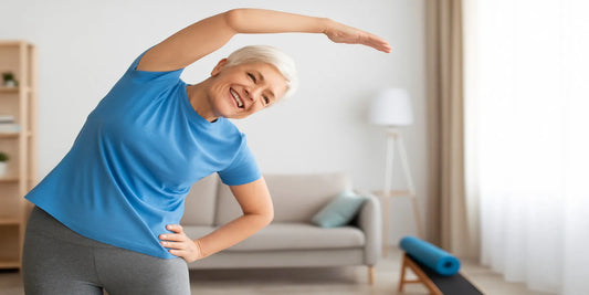 Senior woman with short white hair in blue shirt and gray leggings smiling while stretching indoors during an easy fitness routine.